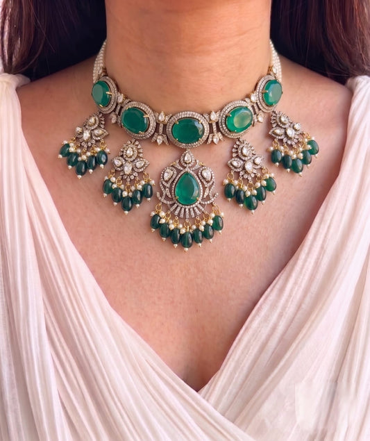 An ornate silver-toned statement necklace with multiple oval and teardrop green gemstones, set with clear stones and dangling green bead chains, worn by a model in a light pink draped top.