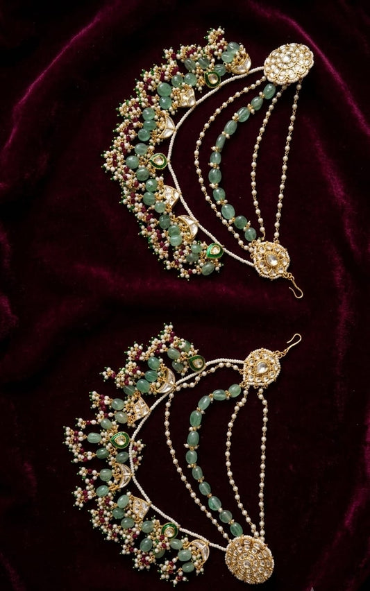 A pair of ornate Indian jhumka earrings with green beads, gold-plated metalwork, and clear stones, displayed on a deep burgundy velvet background.